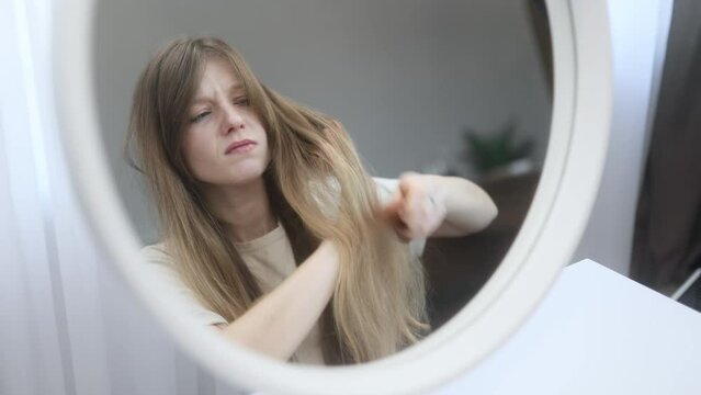 Portrait of nervous caucasian young woman having difficulty brushing her tangled hair dryness split ends problem by wooden brash while looking at mirror reflection at home
