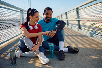 Smiling sports couple using fitness tracker while relaxing outdoors.