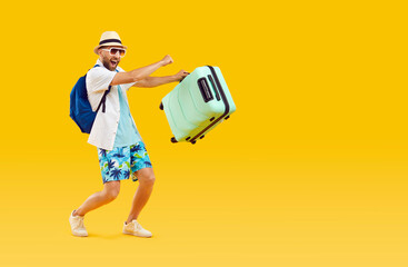 Happy funny male tourist going on summer vacation. Full body length joyful excited young man wearing holiday clothes, with travel suitcase and backpack walking on yellow orange color studio background - obrazy, fototapety, plakaty