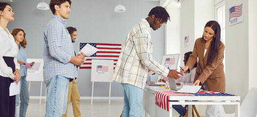 Diverse US citizens perform civic responsibility on election day. African American and Caucasian male and female voters queue up by registration table at voting station in USA. Concept banner - obrazy, fototapety, plakaty