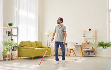 Full length portrait of blind young disabled man in casual clothes with cane walking in the living room at home. Male person with cane indoors. Blindness, disability and vision problems concept.