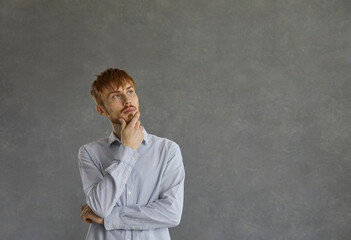 Confident and thoughtful caucasian young man planning his business coming up with an idea. Guy in a shirt holds his hand on his chin and looks thoughtfully to the side standing on a gray background.