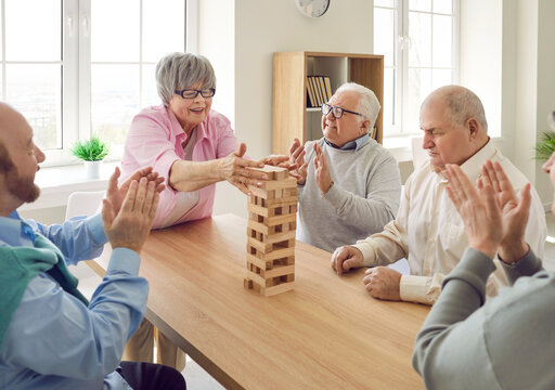 Active old age. Group of senior people sitting at table in nursing home and playing jenga together. Cheerful retired men applauding mature woman who successfully pulls out wooden block in board game.