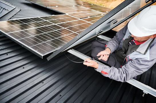 Electrician connecting cables while installing photovoltaic solar panels on roof of house. Worker in white helmet securing cables of solar panel on a roof. Concept of alternative and renewable energy.