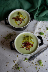 broccoli cream in two black casseroles decorated with a piece of broccoli and ham. They are on a white cloth and pieces of broccoli and a spoon next to a tea towel.