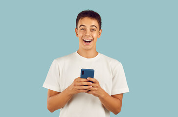 Studio shot of happy young man or adolescent boy in white T shirt isolated on light blue background holding cellphone, reading good news, using mobile phone messenger, looking at camera and smiling