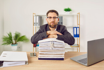 Portrait of a confident young business man accountant in suit working at the desk with a pile of...