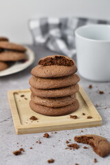 chocolate cookies one on top of the other on a piece of wood. they are on a table with a white cup and a checkered tea towel.