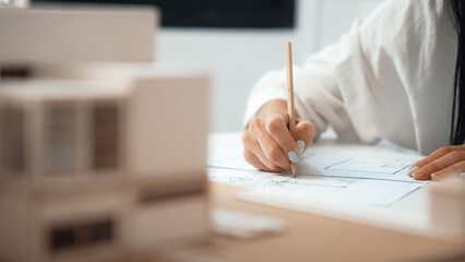 Closeup of beautiful young architect engineer hand drawing, drafting blueprint with blueprint and house model placed on working table at modern office. Focus on hand. Blurring background. Immaculate. - obrazy, fototapety, plakaty