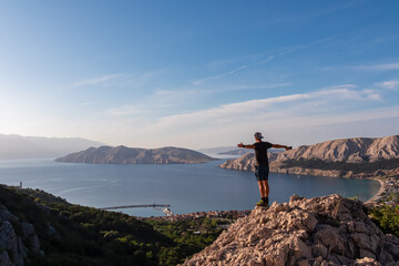Hiker man on edge of rock cliff overlooking archipelago of Kvarner Bay in coastal town Baska, Krk...