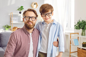 Portrait of a happy young smiling father hugging his cute little child boy wearing eyeglasses. Daddy with son looking cheerful at camera standing at home. Fathers day and fatherhood concept.