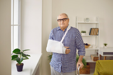 Senior gray-haired elderly man standing at home in rehabilitation with crutch and with arm in...