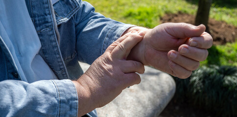 Mature man takes his pulse manually on his wrist while sitting outside on sunny day, close up 
