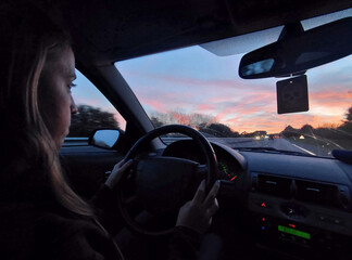 Fototapeta na wymiar Young blonde long-haired girl driving the grand tourer car at sunset on a long trip.Interior view, good driver, attentive and with her hands on the wheel. Dusk with reddish sky