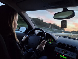 Fototapeta na wymiar Young blonde long-haired girl driving the grand tourer car at sunset on a long trip.Interior view, good driver, attentive and with her hands on the wheel. Dusk with reddish sky