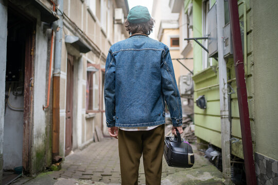 Back view of fashionable young man with vintage cassette player in hand, wandering backstreets of historical city. Trendy modern retro fashion style from 90s.