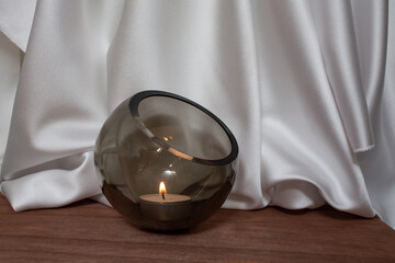 Glass bowl with lit candle on wooden floor with Elegant wavy white satin cloth curtains,  product...