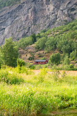 Norwegian fjord mountain scenery with cliffs where there is a red wooden house surrounded by green trees. Sunny summer day.