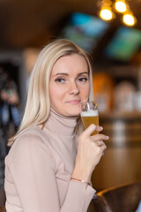 Young cheerful blond woman looking at you while toasting with glass of beer
