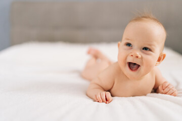 Closeup laughing adorable infant baby lying on bedroom bed, enjoying morning relaxation indoor at home, smiling looking away. Carefree healthy babyhood, healthcare and pediatrics care concept.