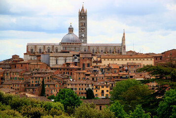 Fototapeta premium View of the historic part of the city of Siena with the Duomo di Siena and green trees and bushes in the foreground against a stormy sky in the Tuscany region of Italy