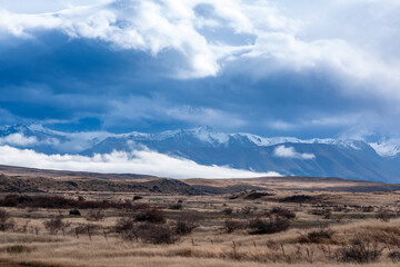 Photograph of a snow-capped mountain range running behind a large brown agricultural field with low level grey clouds on the South Island of New Zealand