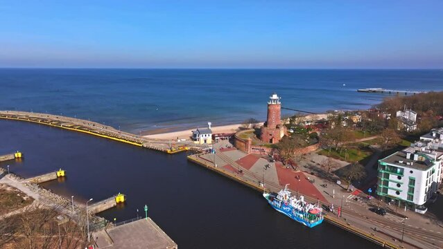 Drone video offers a view of Kolobrzeg port entrance, showcasing a cruise ship and red-brick lighthouse.