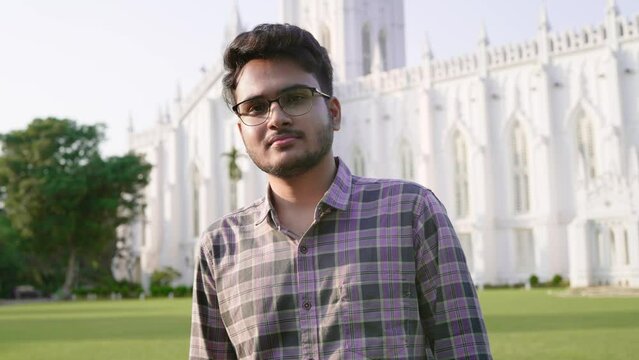 Pan moving shot of a young Indian Asian male or man wearing spectacles is standing outdoors in front of a white building looking or facing the camera smiling on a sunny day.