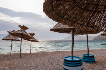 Straw umbrellas at sunrise on idyllic pebble beach in tourist resort town Baska, Krk Otok,...