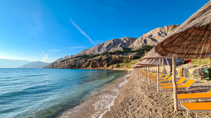 Straw umbrellas and loungers on idyllic pebble beach in resort town Baska, Krk Otok,...