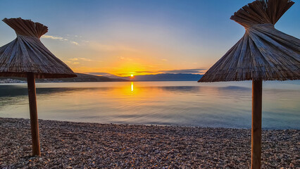 Straw umbrellas at sunset on idyllic pebble beach in tourist resort town Baska, Krk Otok,...