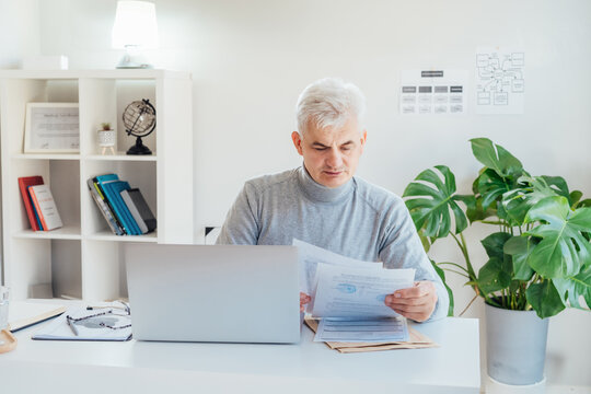 Middle aged businessman reading paper documents and using laptop while working at home office. Confident, experienced senior male professional. Small business entrepreneur manage business paperwork