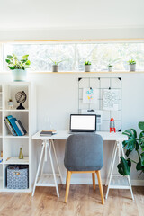 Modern cozy light workplace - white desk with laptop mockup empty screen, grid mood board with pinned notes, shelves with docs and green monstera plant at work space in home office room interior