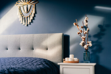 Stylish modern bedroom in dark colors. Cozy interior with navy blue walls, home decor. Bed with gray fabric headboard, blue blanket, bedside table, vase with natural cotton flowers, candles on tray