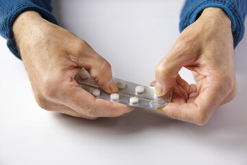 A person is holding a blister of pills and is taking one of them out. The pills are white and the blister is silver. Concept of taking medication and addition to pills.