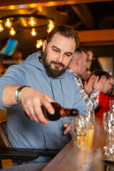 A young handsome man with a beard pouring beer from bottle into glasses