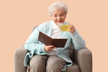 Mature woman with glass cup of lemon tea reading book in armchair on beige background