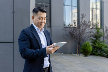 Confident asian businessman smiling while using a tablet outside modern office