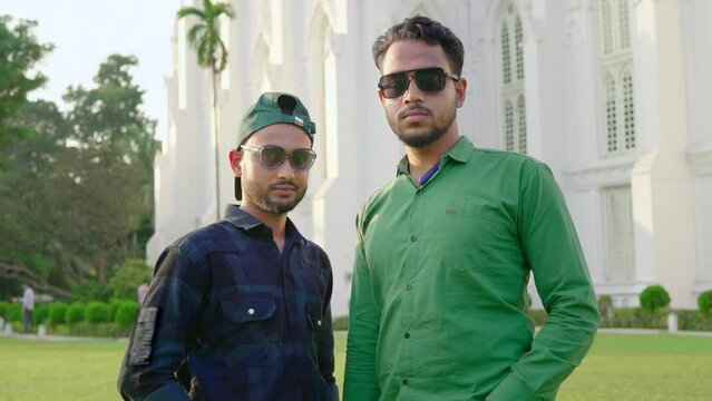 Two young hipster confident Indian Asian men or male friends wearing sunglasses are standing together outdoors in front of the white building looking or facing the camera on a sunny day