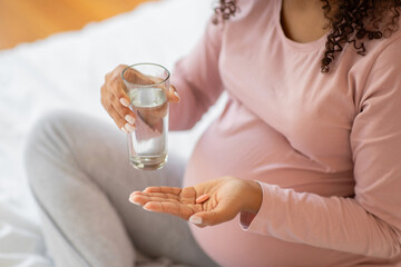 Black pregnant woman taking prenatal vitamins with glass of water at home