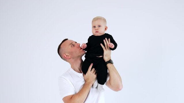 Proud Caucasian dad in white t-shirt is holding a baby on his shoulder. Father and son portrait at white backdrop.