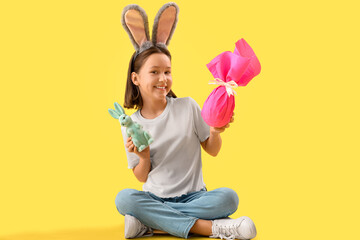 Cute little girl with Easter bunny ears, toy rabbit and gift on yellow background