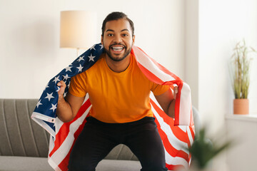 Emotional African American soccer fan, jumping off sofa at home with USA flag for goal celebration