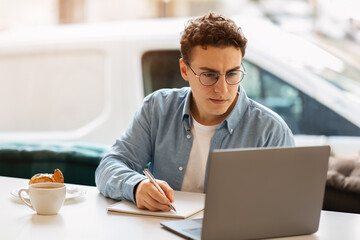 A studious young man with curly hair and glasses is writing in a notebook at a cafe table with a...