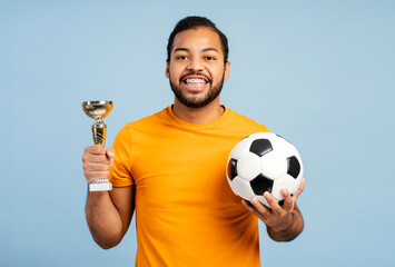 African American athlete with braces holding championship cup and soccer ball, looking at the camera