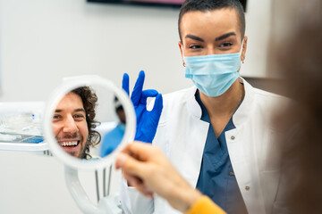 A happy man looking himself in the hand held mirror at the dentist's office. Female dentist showing...