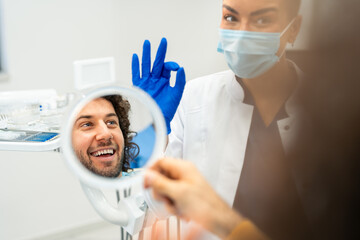 Female dentist showing a hand sign for perfection. Confident man with a beautiful healthy smile holding a vanity mirror.