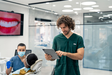 Obraz premium Handsome male dentist in green uniform working on digital tablet in modern dental office with patient in dentist's chair and nurse in background.