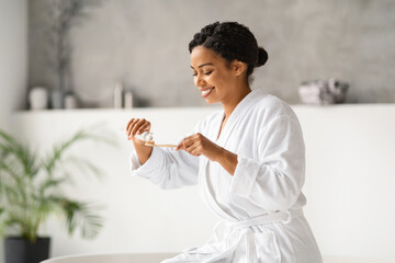 Happy black woman in bathrobe preparing to brush her teeth, applying toothpaste