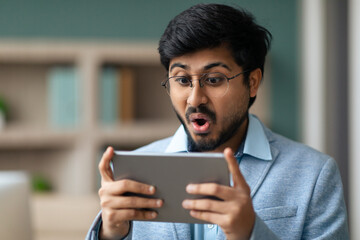 Excited Hindu entrepreneur guy using digital tablet at modern workplace